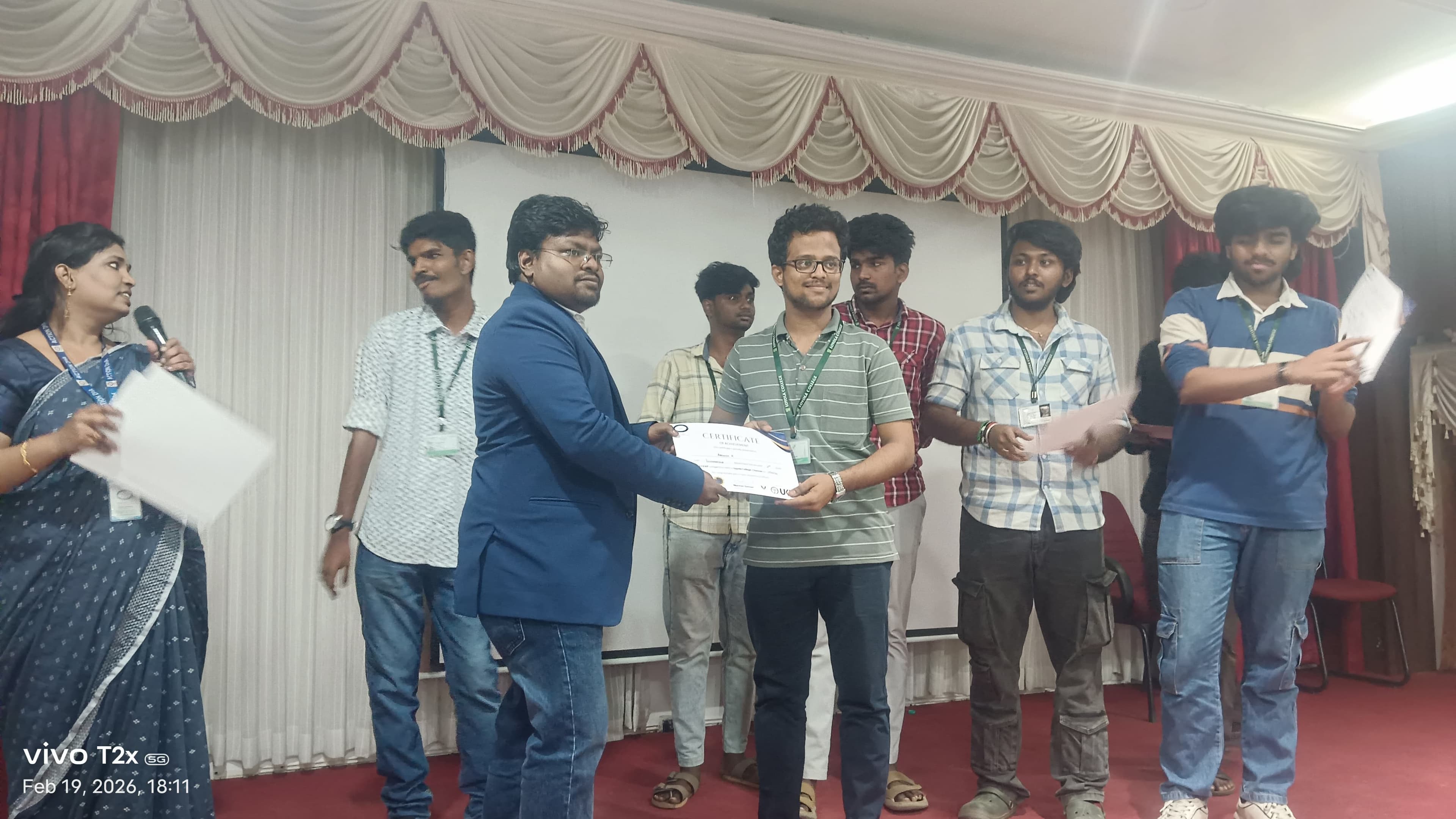 Facilitator presenting a certificate to a student on a college auditorium stage